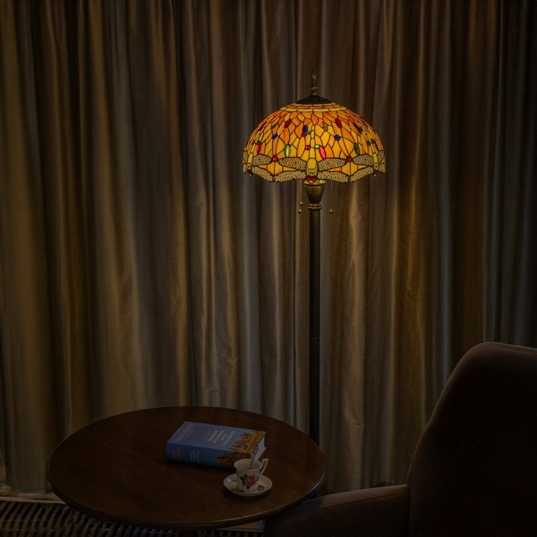 Een klassieke Tiffany vloerlamp met een warmgekleurd glas-in-lood kap in rood, geel en blauw tinten, verlicht een gezellige zithoek met een houten tafel, een boek en een theekopje erop, voor gesloten gordijnen.
