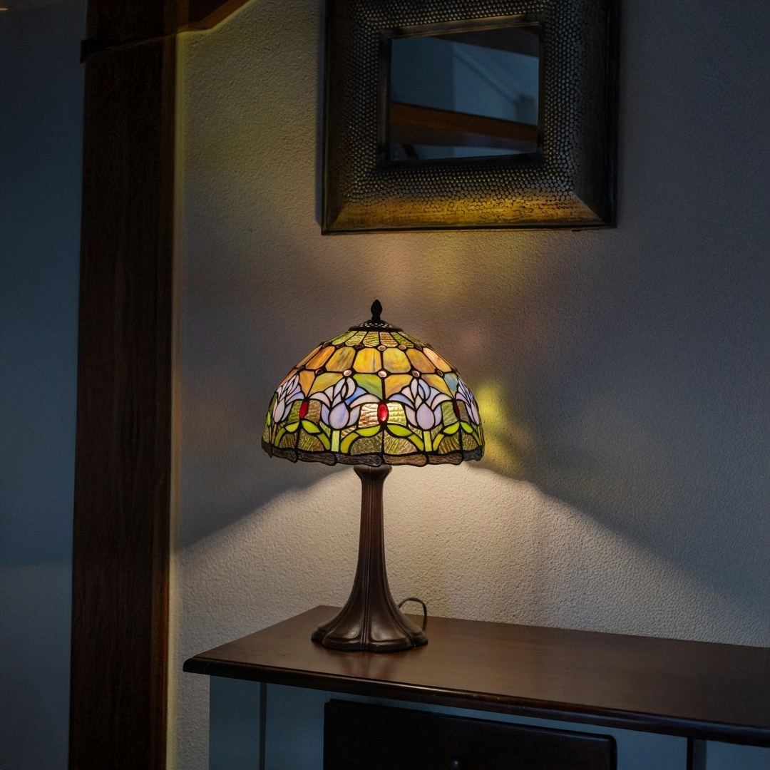 Een Tiffany tafellamp met een bronzen voet en een kleurrijke glas-in-lood kap met witte bloemen en groene bladeren, geplaatst op een houten kast tegen een lichte muur, met een spiegel erboven.