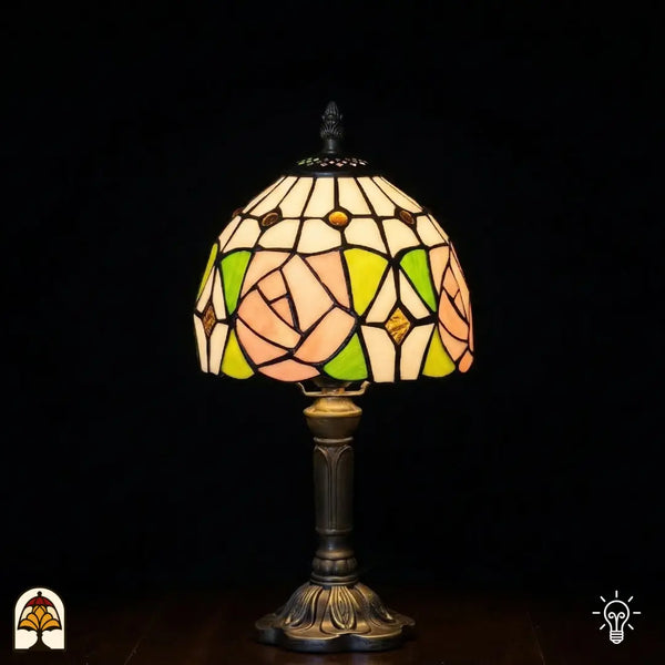 Verlichte kleine Tiffany tafellamp Rosalia Verde met koepelvormige kap, grote roze roos en groene bladeren in glas-in-lood, witte bovenkap met gouden ronde accenten, bronzen geribbelde vaasvoet, donkere achtergrond