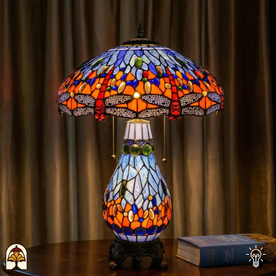 Tiffany lamp tafellamp verlicht op houten tafel met gordijn op de achtergrond, blauw-oranje kap met libellemotieven en blauwe glaskralen, lichtblauwe glas-in-lood peervormige voet en bronzen leeuwenpootvoet