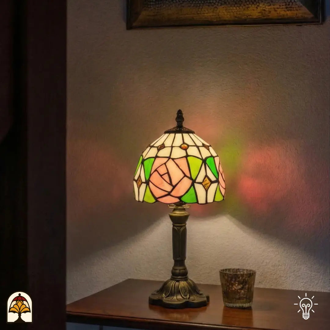 Verlichte Tiffany tafellamp Rosalia Verde op houten dressoir naast een sfeerlichtje, roze roos en groene bladeren verlicht, kleurrijke lichtreflecties op muur