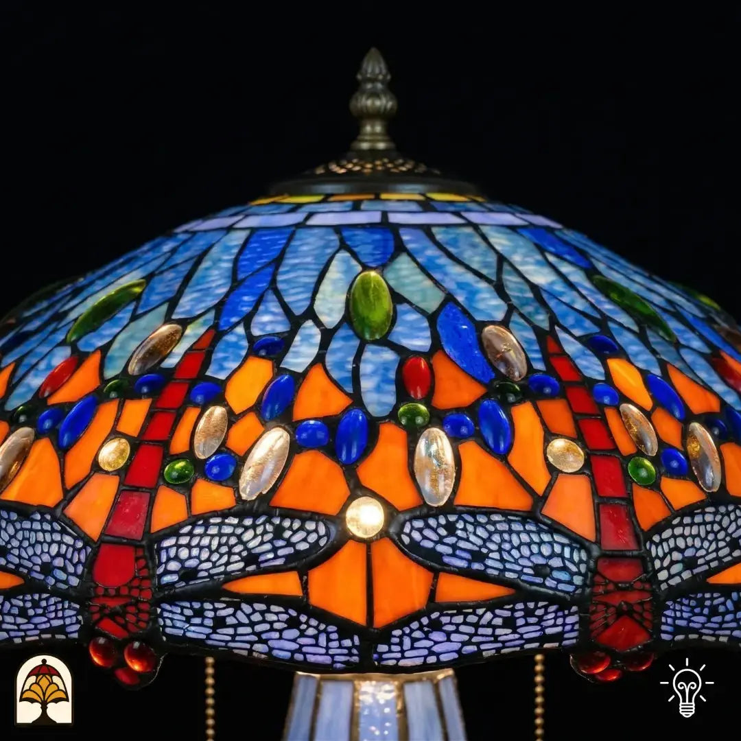 Detailopname van tiffany lamp kapbovenzone verlicht, blauw en lichtblauw glas met blauwe, groene en amber ovale glaskralen in een band, puntfinial bovenaan zichtbaar