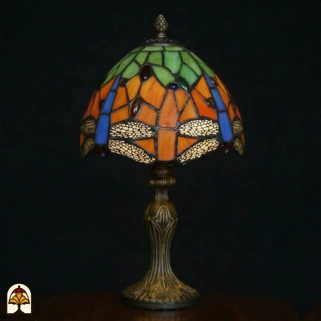 Onverlichte Tiffany tafellamp Libellula Azul ⌀20 op donkere achtergrond, oranje kap met groene bovenpanelen, blauwe libellenvleugels en libellenfries, rode en groene juwelen, bronzen Art Nouveau voet