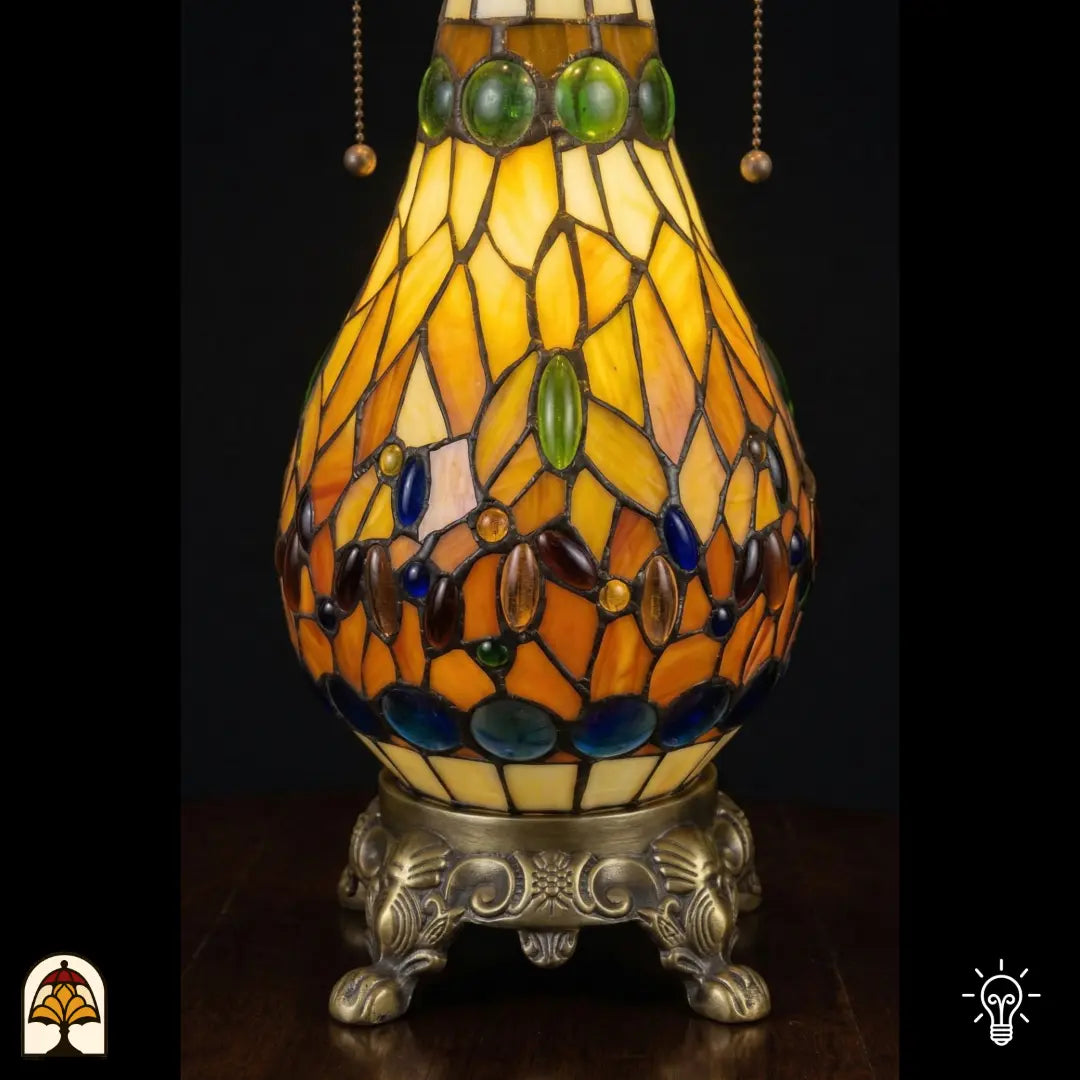 Detailopname van tiffany lamp glas-in-lood peervormige voet verlicht, amberkleurig glas met groene, blauwe en bruine ovale glaskralen, bronzen siervoet met leeuwenpoten en acanthusrolwerk
