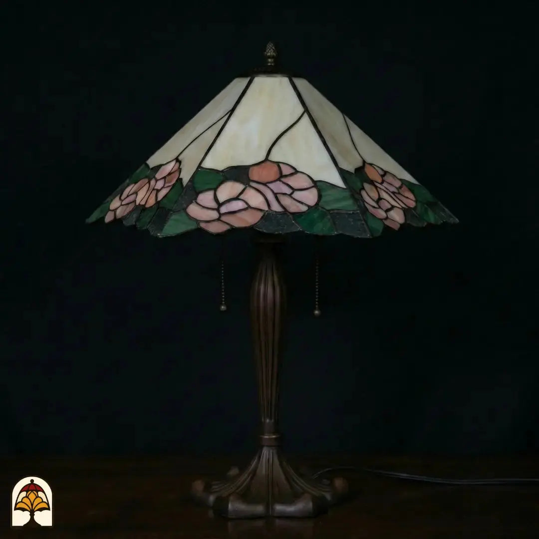 Onverlichte Tiffany tafellamp Alba Sereno op donkerblauwe achtergrond, brede trapeziumvormige kap met roze rozen en groene bladeren, bronzen voet met trekschakelaars