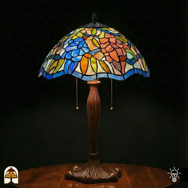 Verlichte Tiffany tafellamp Florentia Blu met brede koepelvormige kap, blauwe bloemen en oranje-roze bladmotieven in glas-in-lood, blauwe rand onderaan, bronzen geribbelde vaasvoet met trekschakelaars, donkere achtergrond