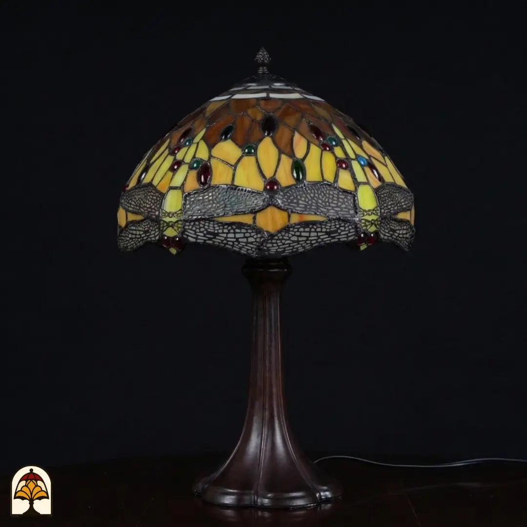 Onverlichte Tiffany tafellamp Libellula Aurea ⌀30 op donkere achtergrond, koepelvormige kap met amber-bruine en gele panelen, libellenfries met grijze vleugels, rode en groene juwelen, bronzen trompetvoet