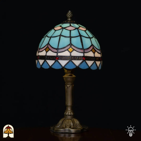 Verlichte Tiffany tafellamp Sapphire ⌀20 cm op zwarte achtergrond – lichtblauwe koepelkap met visschubbenpatroon, roze sierband, rode juwelen en bronzen voet