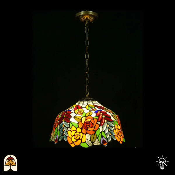 Tiffany lamp hanglamp verlicht, koepelvormige glas-in-lood kap met oranje-gele rozen, rode ovale glaskralen en groene bladeren op witte achtergrond, gekartelde rand, opgehangen aan bronzen ketting met sierlijke plafondrozet
