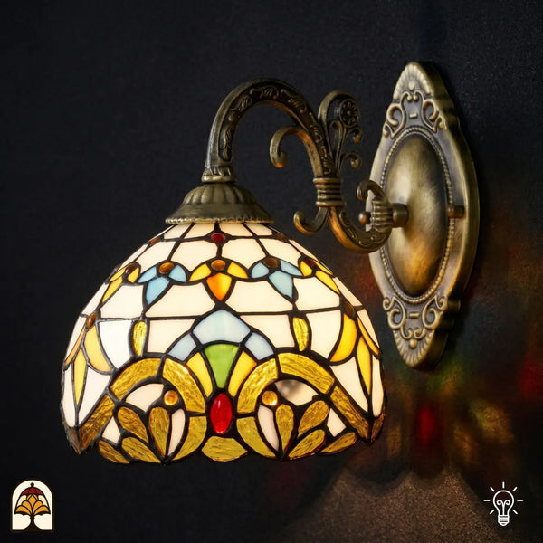 Verlichte Tiffany wandlamp Barok vooraanzicht met goudgele glas-in-lood kap, blauwe en groene accenten, op bronzen gebogen arm met ovale wandplaat