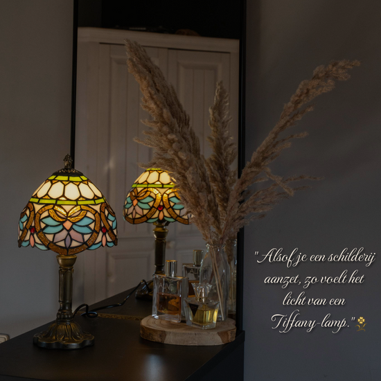 Sierlijke Tiffany tafellamp met bloemmotief in pastel- en goudtinten, geplaatst op een dressoir met spiegel en droogbloemen. Handgemaakte lamp die zorgt voor een warme, nostalgische gloed.