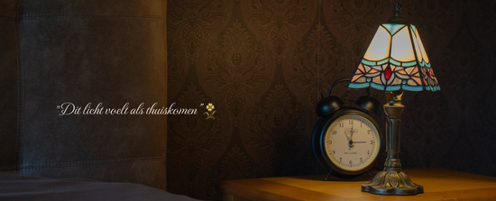 Tiffany tafellamp met elegante geometrische kap in zachte crème- en blauwtinten, staand op een nachtkastje met wekker. Perfecte sfeerlamp die een huiselijke en klassieke gloed verspreidt.