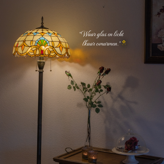 Statige Tiffany vloerlamp met royale kap in geel- en blauwtinten, staand naast een houten bijzettafel met rozen en kaarsjes. Klassieke handgemaakte vloerlamp die de woonkamer sfeervol verlicht.