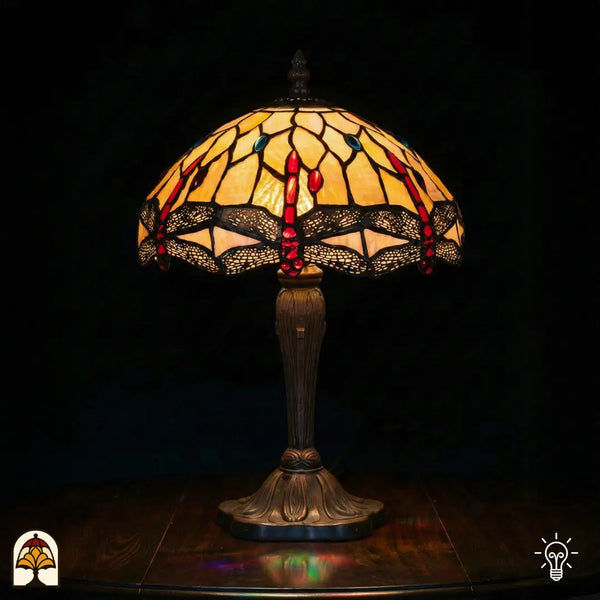Verlichte Tiffany tafellamp Libelle Elegance met koepelvormige geel-amber kap, rode druppelvormige en blauwe ronde glas-in-lood accenten, libellenmotieven langs de rand, bronzen geribbelde vaasvoet, donkere achtergrond