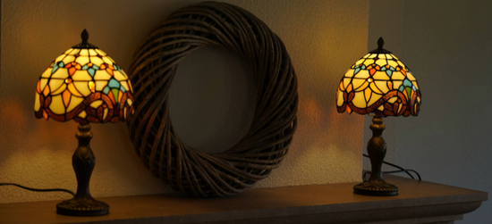 Twee brandende Tiffany-stijl tafellampen met glas-in-lood kappen in warme geel-, oranje- en blauwtinten, geplaatst op de bovenkant van een open haard met daarachter een grote decoratieve rotan krans aan de muur.