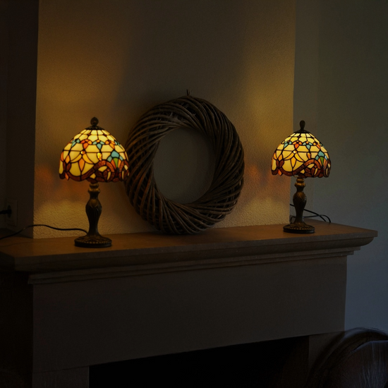 "Twee brandende Tiffany-stijl tafellampen met glas-in-lood kappen in warme geel-, oranje- en blauwtinten, geplaatst op de bovenkant van een open haard met daarachter een grote decoratieve rotan krans aan de muur.