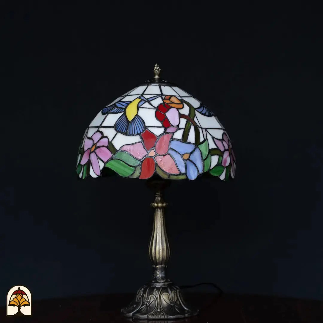Tiffany tafellamp Colibrì onverlicht op zwarte achtergrond — wit/crème kap met kolibrie en bloemen in roze, rood, blauw en groen, bronzen kolomvoet