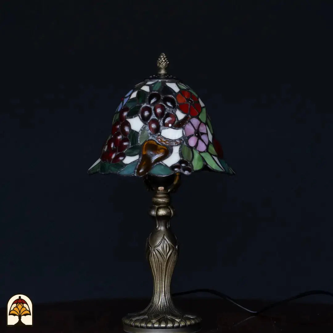 Onverlichte Tiffany tafellamp op donkere achtergrond — klokvormige kap met bordeaux druiven, amber glas, roze en blauwe bloemen, groene bladeren, klassieke kandelaarvoet