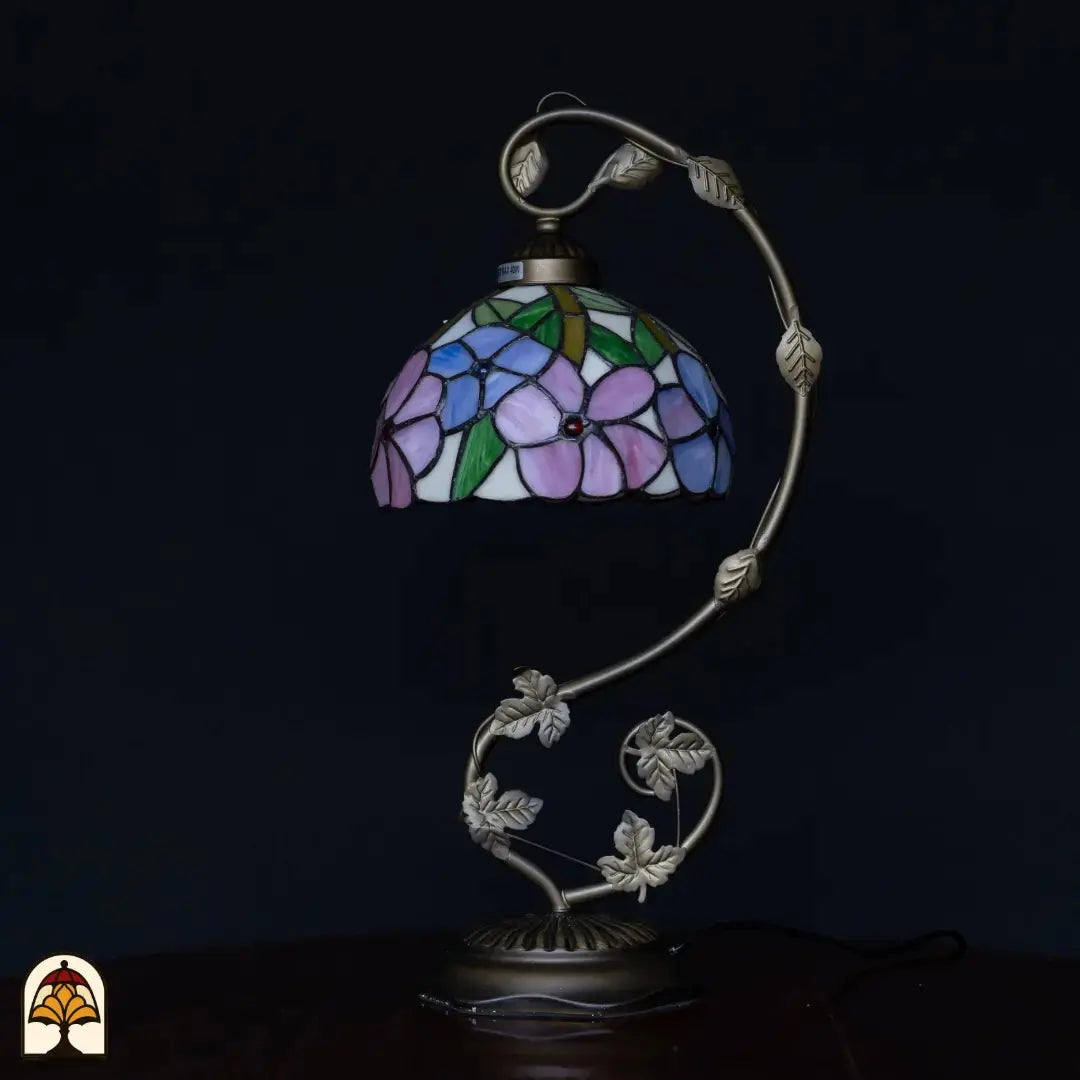 Onverlichte Tiffany tafellamp op donkere achtergrond — kap met blauw/paarse en roze bloemen, groene bladeren, zilveren gebogen arm met blad-details