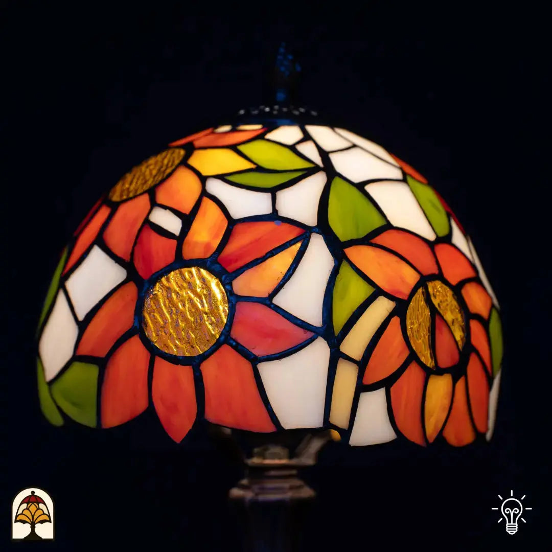 Close-up verlichte kap Tiffany tafellamp Solea — oranje zonnebloembladen met goud/bruine centra, groene bladeren, crème/witte vlakken