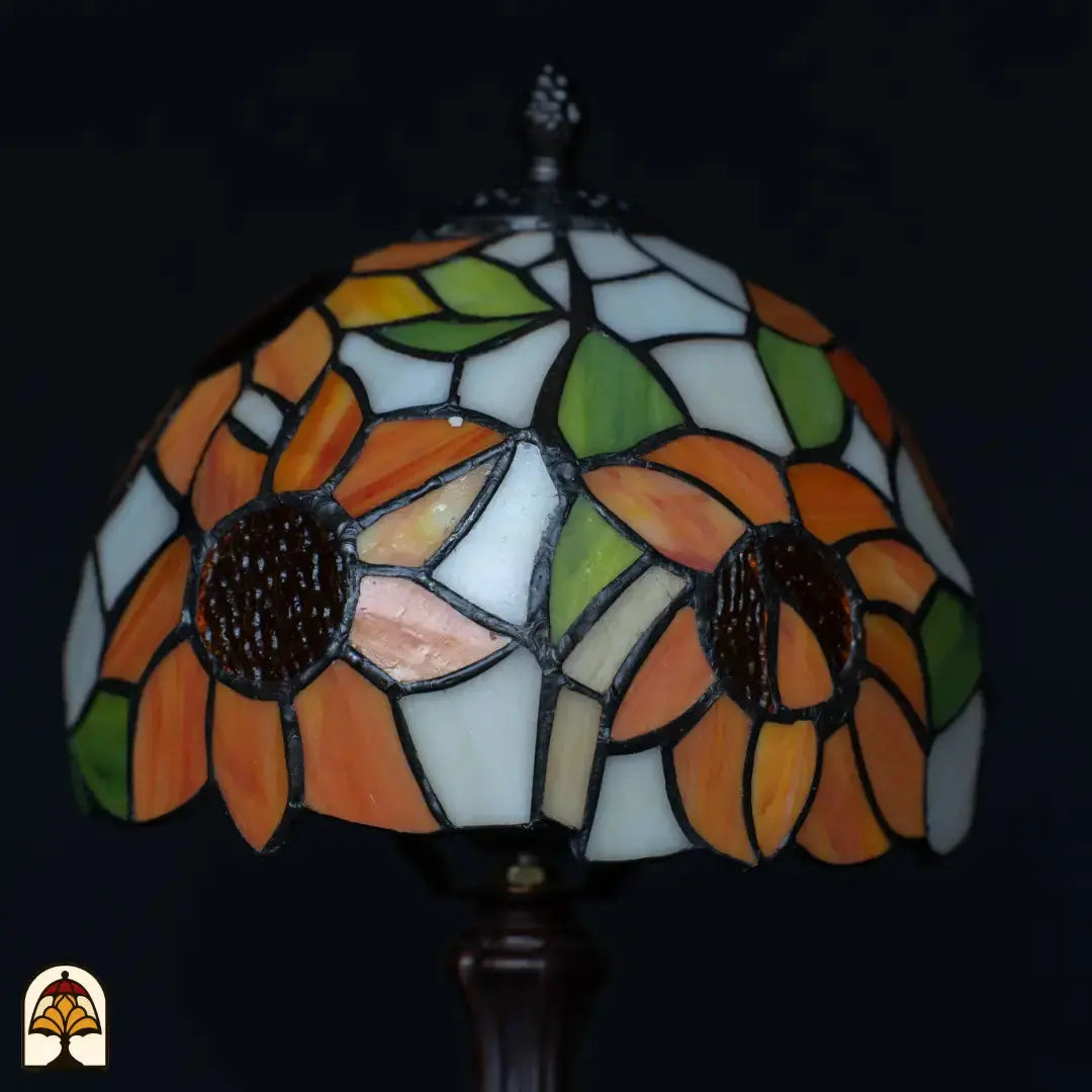 Close-up kap Tiffany tafellamp Solea onverlicht — oranje zonnebloembladen met donkerbruine centra, crème/witte en blauwe vlakken, groene bladeren