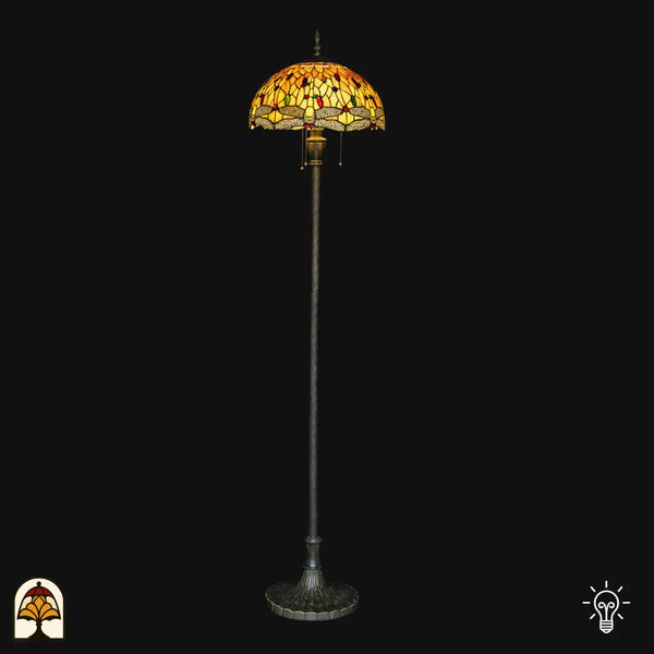 Tiffany vloerlamp Libellula Aurea verlicht op zwarte achtergrond — amber/gele koepelkap met libellenrand, lange paal met brede bronzen voet, trekschakelaars