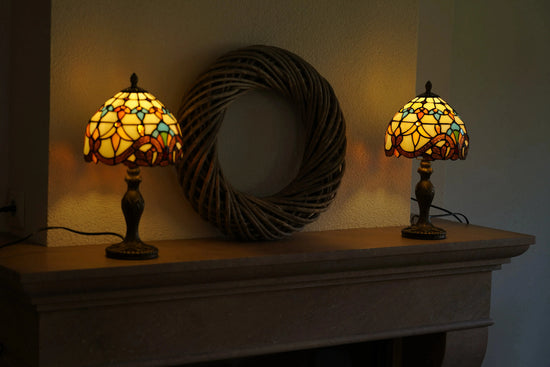 Twee brandende Tiffany tafellampen met warm geel, oranje en turquoise glas staan symmetrisch op een stenen schouw, met een grote ronde rieten wanddecoratie ertussen. De lampen verspreiden zacht, sfeervol licht tegen de wand.