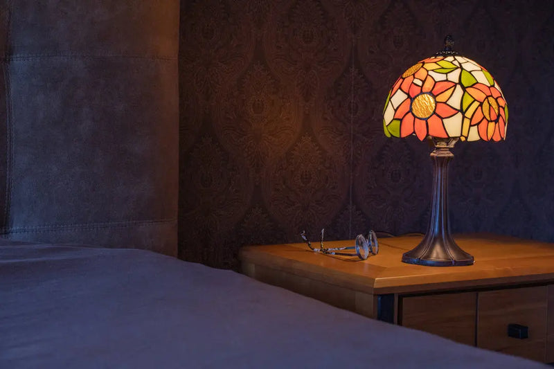 Een verlichte, decoratieve Tiffany-stijl glas-in-loodlamp met oranje en gele bloemenpatronen staat op een houten nachtkastje. Een bril rust ernaast. Op de voorgrond bevindt zich de hoek van een bed met paars beddengoed, tegen een donker damastbehang.