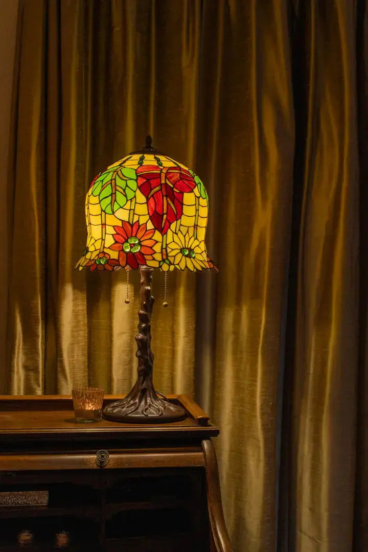 Tiffany tafellamp met een glas-in-lood kap vol bloem- en bladvormen in warme tinten rood, geel en groen. De lamp straalt een zacht, nostalgisch licht uit op een houten bureau voor goudkleurige gordijnen.