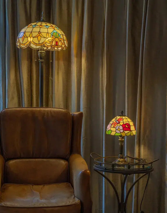 Tiffany vloerlamp en tafellamp in een warme woonkamerhoek, naast een bruine leren fauteuil en een glazen bijzettafel, beide lampen met glas-in-lood kappen die zacht licht uitstralen tegen een achtergrond van beige gordijnen.