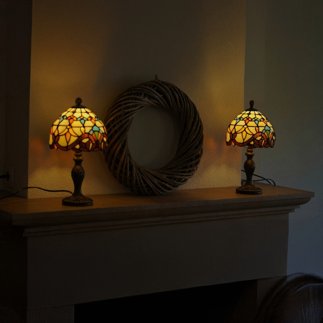 Twee brandende Tiffany-stijl tafellampen met glas-in-lood kappen in warme geel-, oranje- en blauwtinten, geplaatst op de bovenkant van een open haard met daarachter een grote decoratieve rotan krans aan de muur.