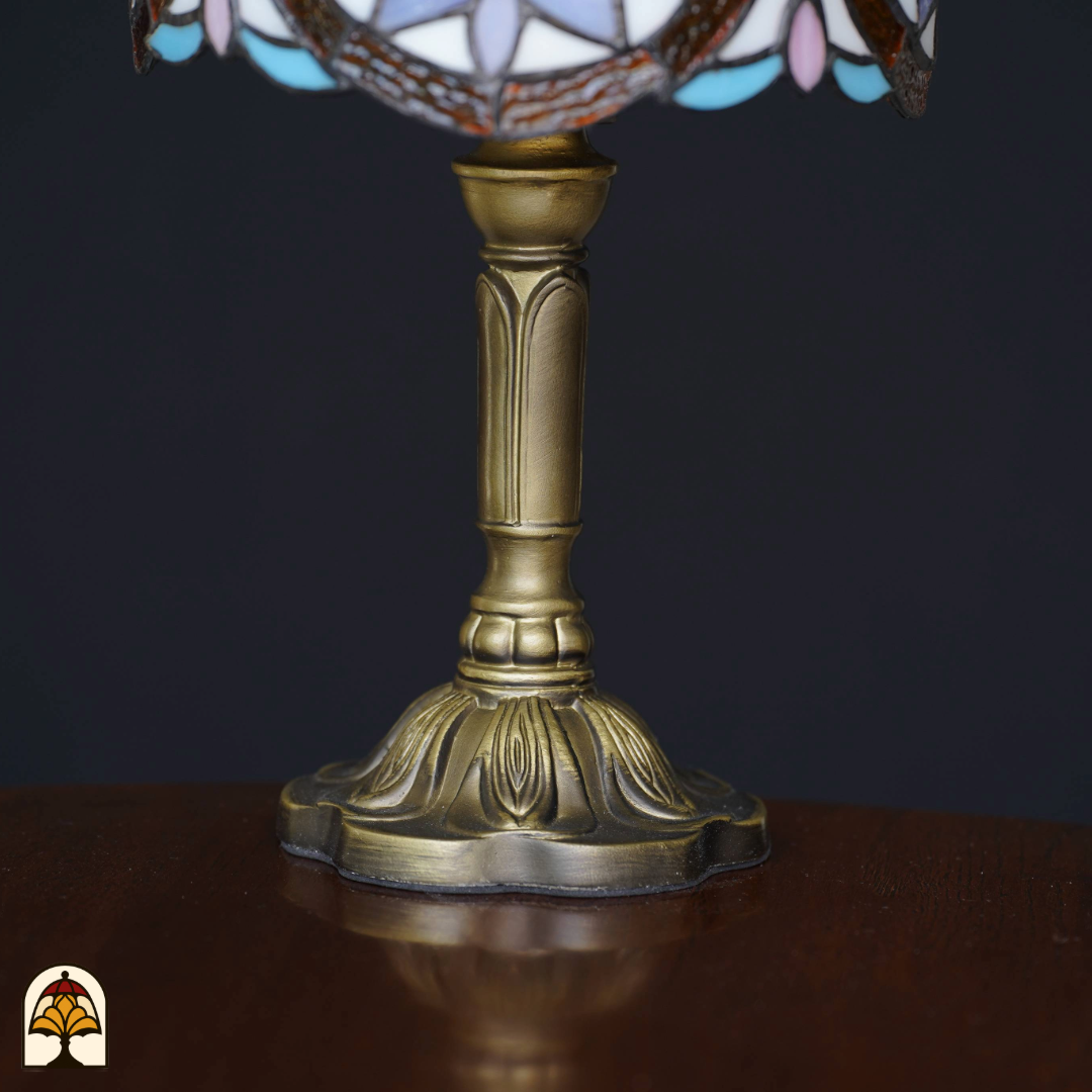 Close-up van de sierlijke bronzen voet van een Tiffany-stijl tafellamp, geplaatst op een houten tafel tegen een zwarte achtergrond. De lamp staat uit.