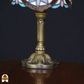 Close-up van de sierlijke bronzen voet van een Tiffany-stijl tafellamp, geplaatst op een houten tafel tegen een zwarte achtergrond. De lamp staat uit.