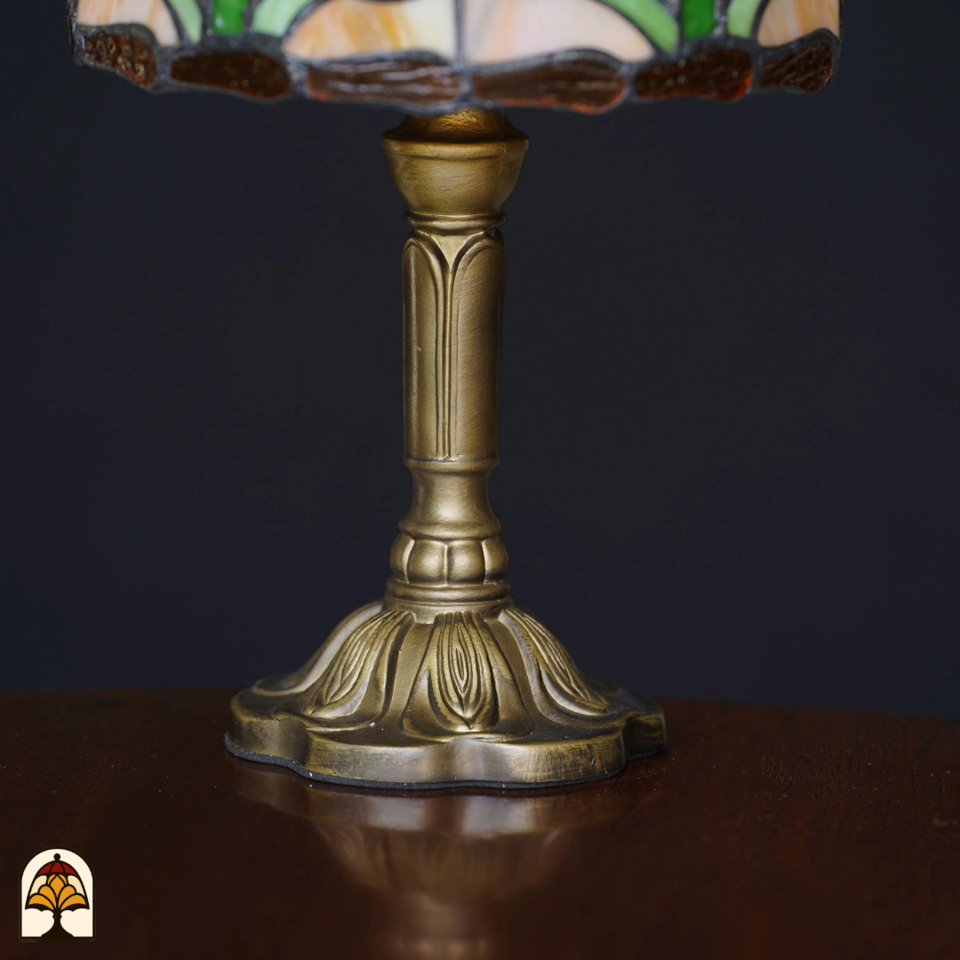 Close-up van de sierlijke bronzen voet van een Tiffany-stijl tafellamp, geplaatst op een houten tafel tegen een zwarte achtergrond. De lamp staat uit.
