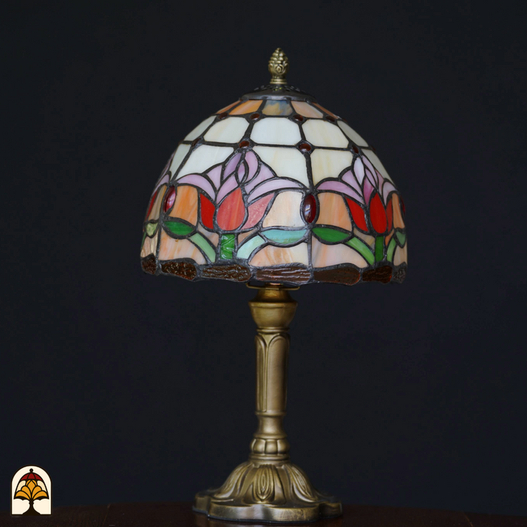 Close-up van een Tiffany-stijl lampenkap met rood-, roze-, groen- en witkleurige tulpenmotieven, tegen een zwarte achtergrond. De lamp staat uit.