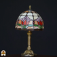 Close-up van een Tiffany-stijl lampenkap met rood-, roze-, groen- en witkleurige tulpenmotieven, tegen een zwarte achtergrond. De lamp staat uit.