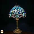 Onverlichte Tiffany tafellamp Libellula Celeste ⌀20 op donkere achtergrond, koepelvormige kap met lichtblauwe en turquoise glas-in-lood panelen, witte ovale juwelen, libellenfries onderaan, bronzen gedraaide steel met bladrelief voetplaat