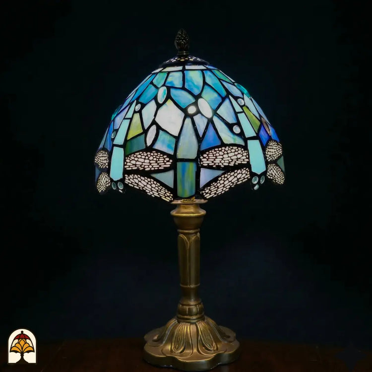 Onverlichte Tiffany tafellamp Libellula Celeste ⌀20 op donkere achtergrond, koepelvormige kap met lichtblauwe en turquoise glas-in-lood panelen, witte ovale juwelen, libellenfries onderaan, bronzen gedraaide steel met bladrelief voetplaat