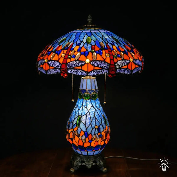 Tiffany lamp tafellamp verlicht, koepelvormige kap met blauw glas-in-lood bovenzone, oranje-rode middenzone met blauwe en amber glaskralen, lavendelblauwe libelle rand onderaan; peervormige lichtblauwe voet en bronzen leeuwenpootvoet
