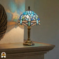 Verlichte Tiffany tafellamp Libellula Celeste ⌀20 op schoorsteenmantel in interieur, blauw licht gereflecteerd op witte wand, bronzen voet zichtbaar
