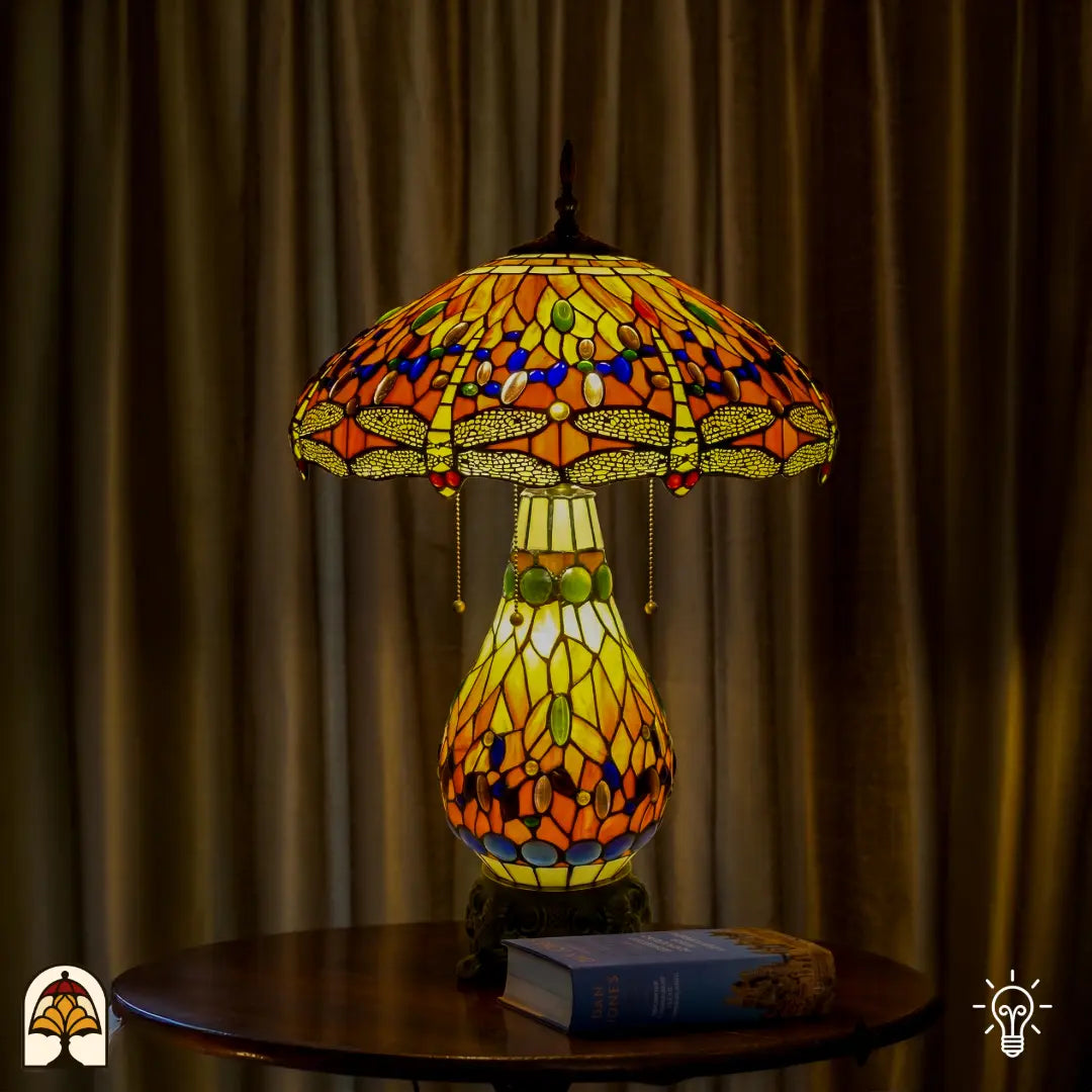 Tiffany lamp tafellamp verlicht op houten tafel met gordijn op de achtergrond, amberkleurige kap met libellemotieven en blauwe glaskralen, glas-in-lood peervormige voet en bronzen leeuwenpootvoet