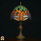 Onverlichte Tiffany tafellamp Libellula Azul ⌀20 op donkere achtergrond, oranje kap met groene bovenpanelen, blauwe libellenvleugels en libellenfries, rode en groene juwelen, bronzen Art Nouveau voet