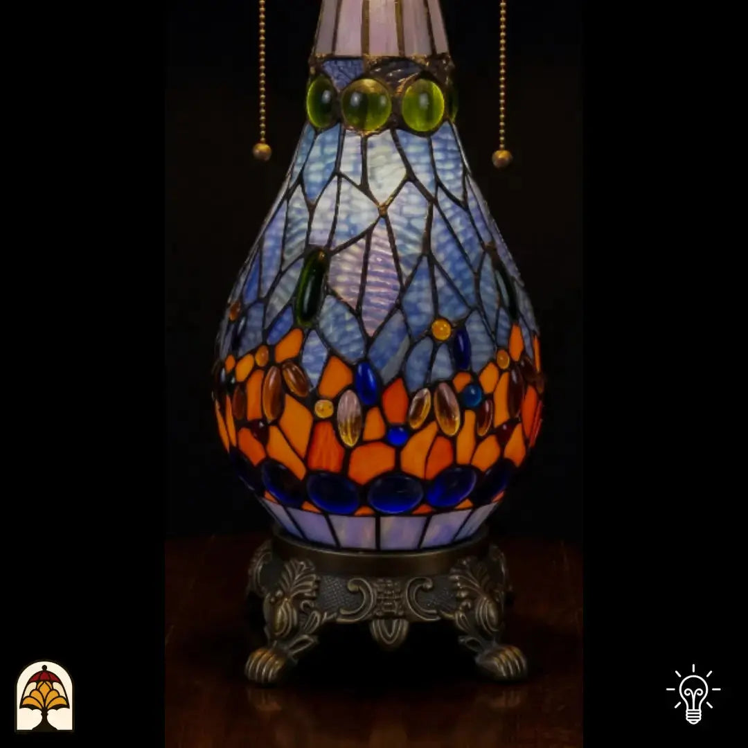 Detailopname van tiffany lamp glas-in-lood peervormige voet verlicht, lichtblauw glas met groene, blauwe en oranje glaskralen, trekketting zichtbaar en bronzen siervoet met leeuwenpoten