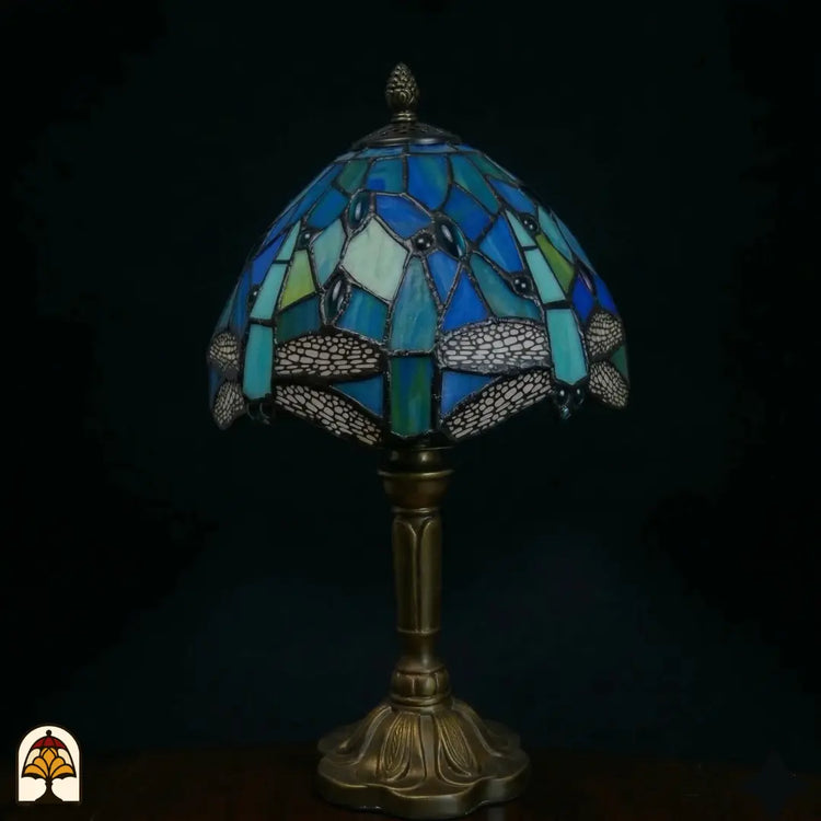 Onverlichte Tiffany tafellamp Libellula Celeste ⌀20 op donkere achtergrond, kap met donkerblauwe en turquoise glas-in-lood panelen, libellenfries, bronzen steel met bloemenrelief voet