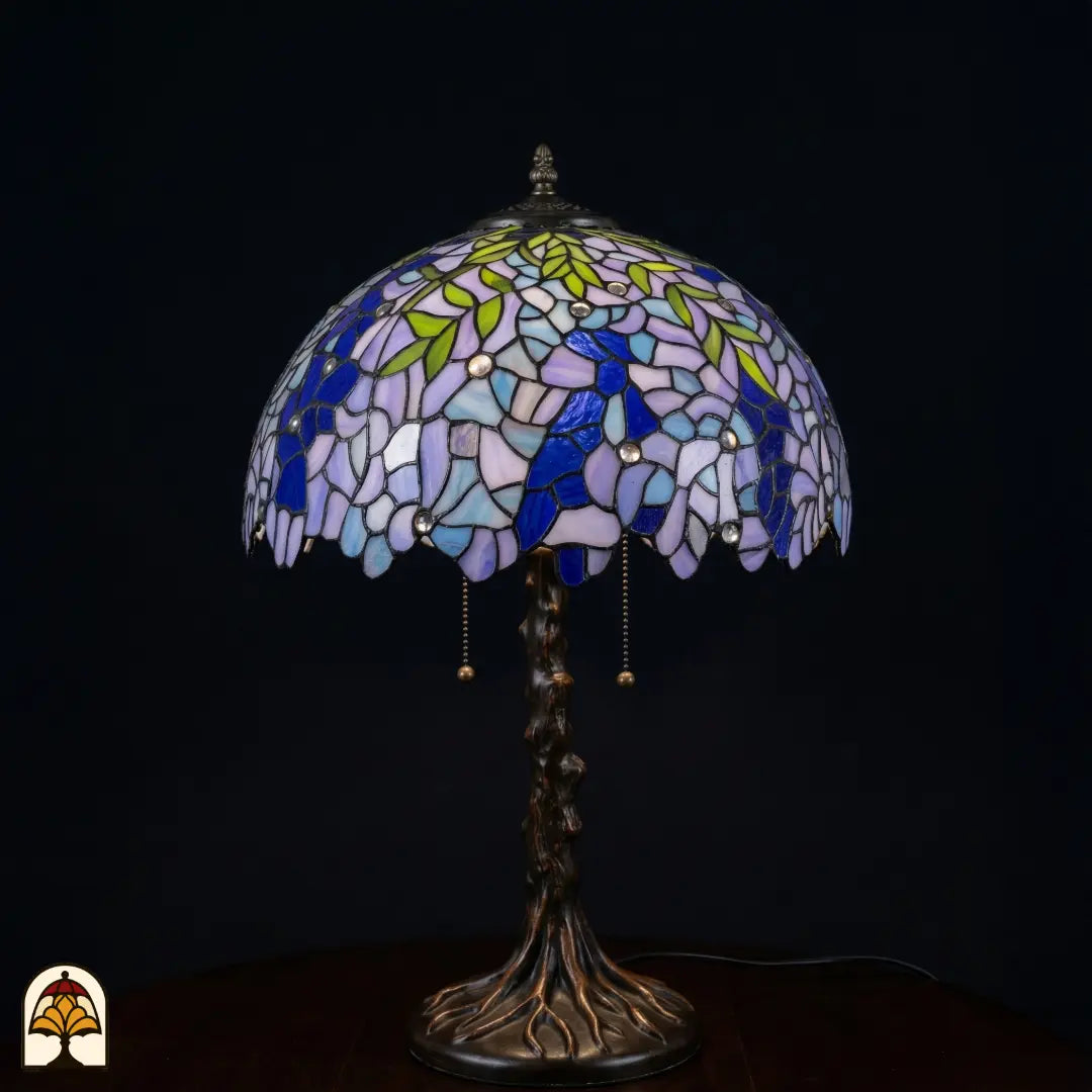 Onverlichte Tiffany tafellamp Wisteria Celeste op donkere achtergrond met lila-blauwe en witte glyciniabloemen, groene bladeren en bronzen boomstam voet met wortels
