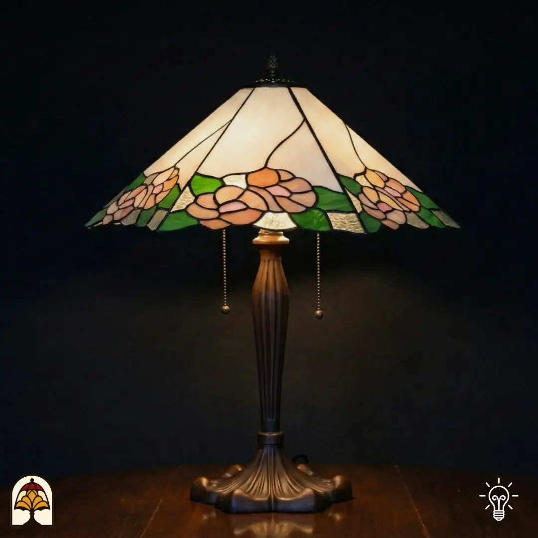 Verlichte Tiffany tafellamp Alba Sereno met brede trapeziumvormige kap, witte en crème bovenkap met roze rozen en groene bladeren langs de rand, bronzen geribbelde voet met trekschakelaars, donkere achtergrond