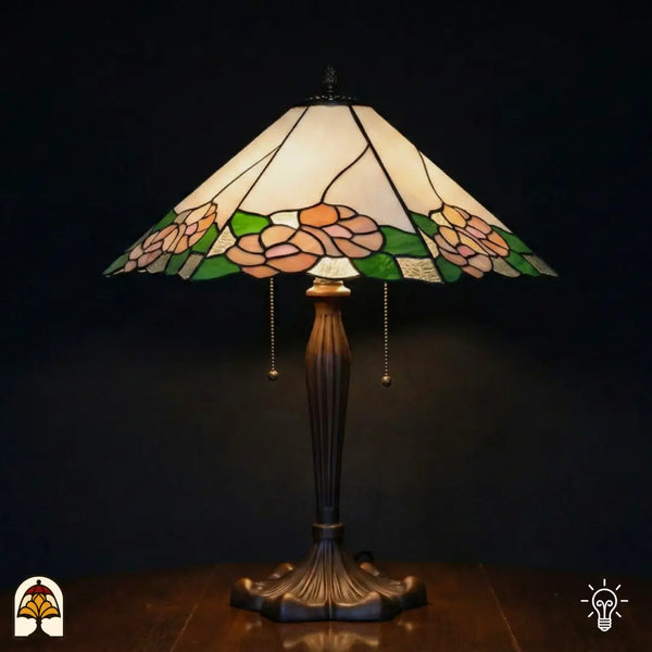 Verlichte Tiffany tafellamp Alba Sereno met brede trapeziumvormige kap, witte en crème bovenkap met roze rozen en groene bladeren langs de rand, bronzen geribbelde voet met trekschakelaars, donkere achtergrond