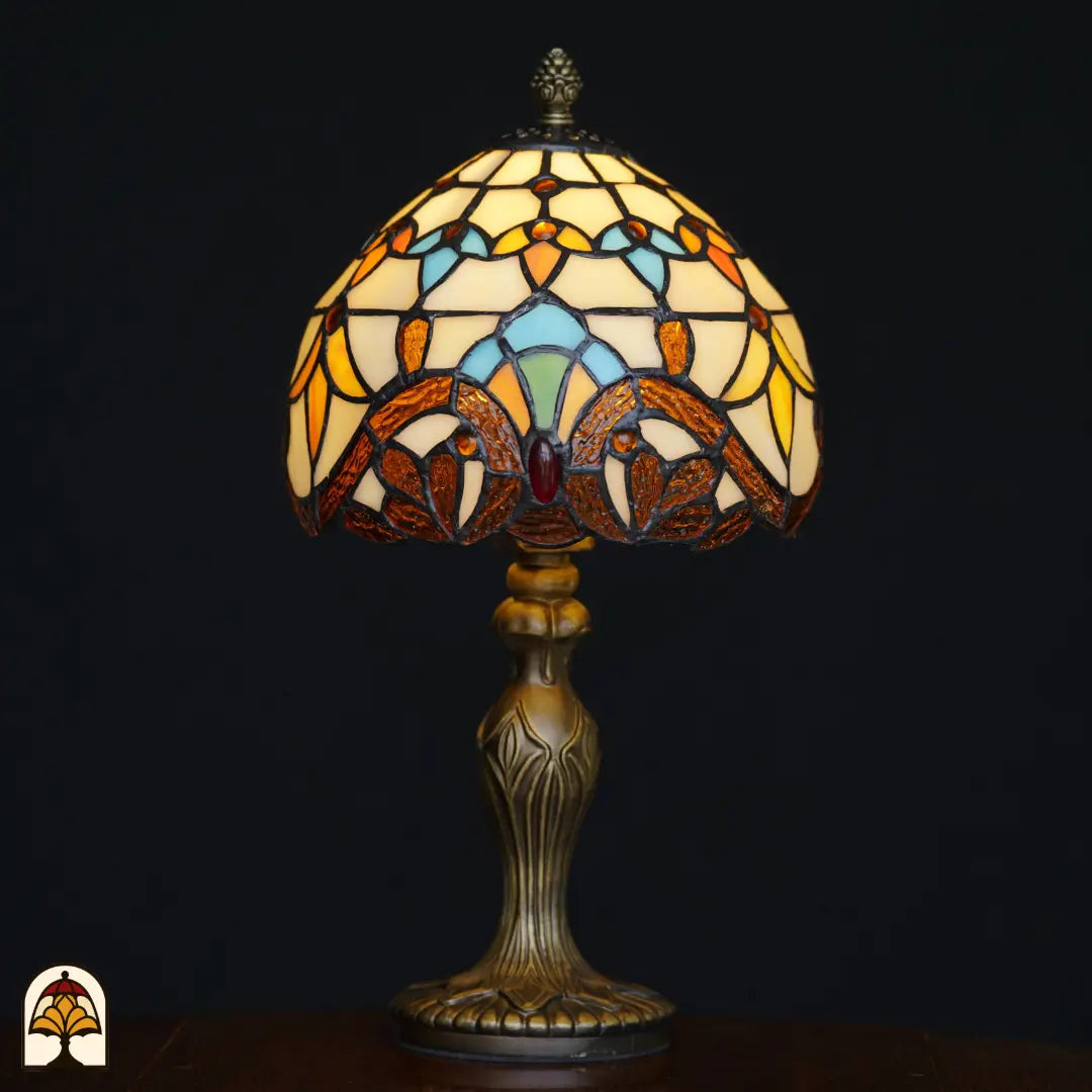 Verlichte Tiffany tafellamp Baroque ⌀20 cm op zwarte achtergrond – crème koepelkap met amber barokke bladvormen, lichtblauwe boogvormen en rode edelsteen, sierlijke bronzen Art Nouveau voet
