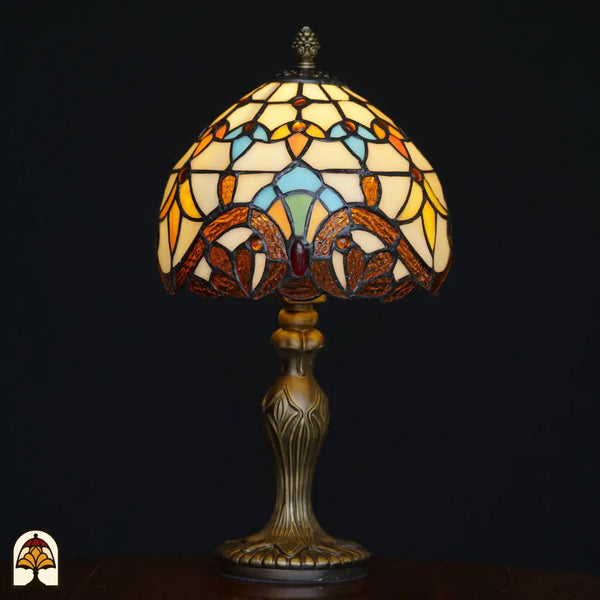 Verlichte Tiffany tafellamp Baroque ⌀20 cm op zwarte achtergrond – crème koepelkap met amber barokke bladvormen, lichtblauwe boogvormen en rode edelsteen, sierlijke bronzen Art Nouveau voet