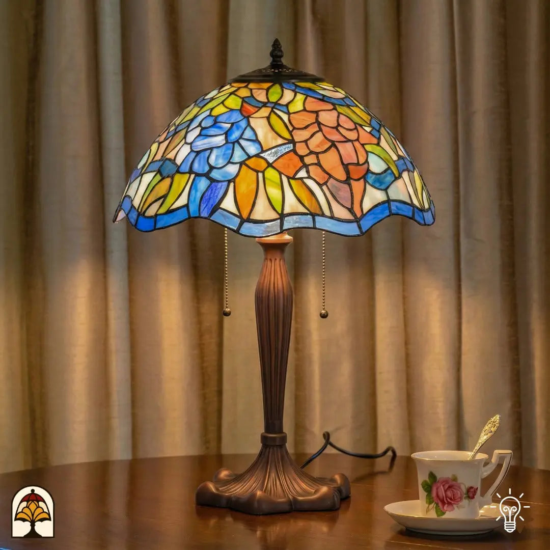 Verlichte Tiffany tafellamp Florentia Blu op houten tafel voor gordijnen, blauwe en oranje bladmotieven verlicht, theekopje met schotel ernaast